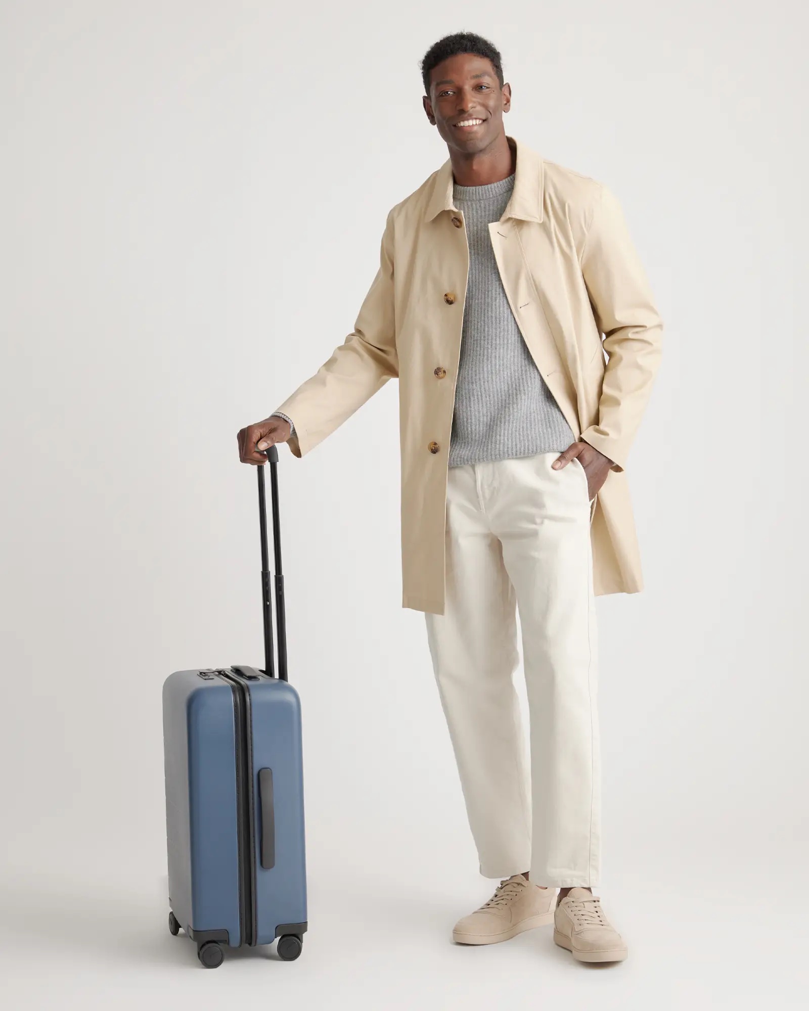 A man in a trench coat holds the hand le of a blue carry on case. 