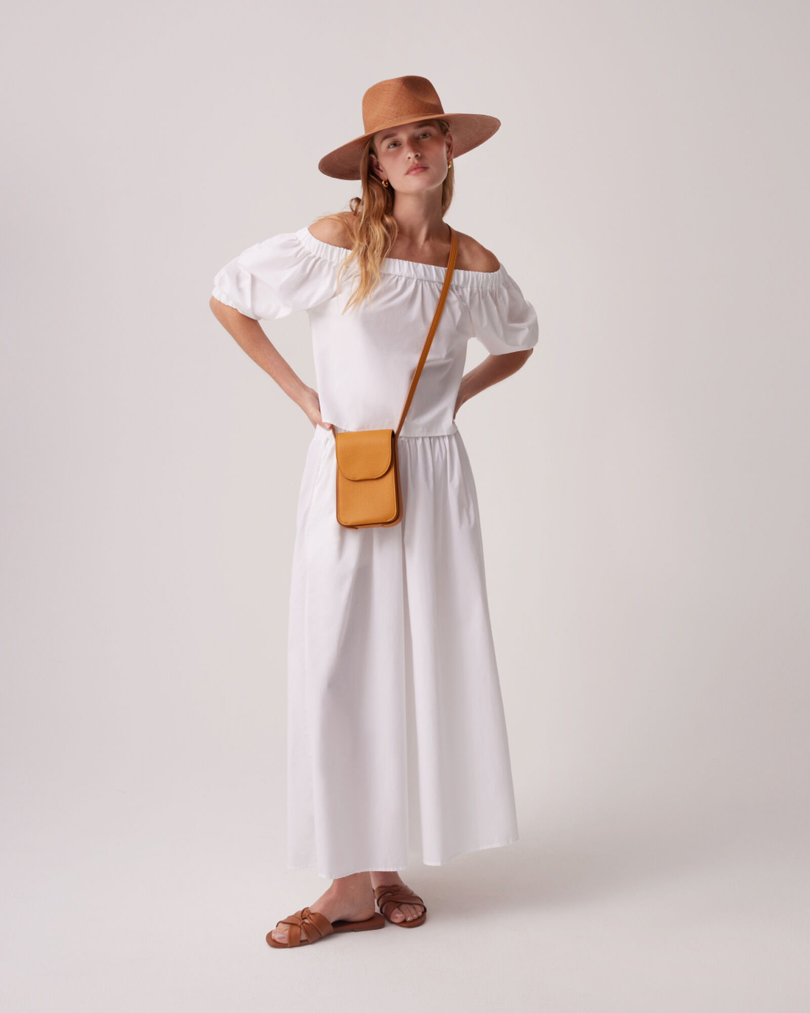 A model wearing a fedora and off the shoulder white top with matching white flowy pants and a yellow leather cell phone bag.