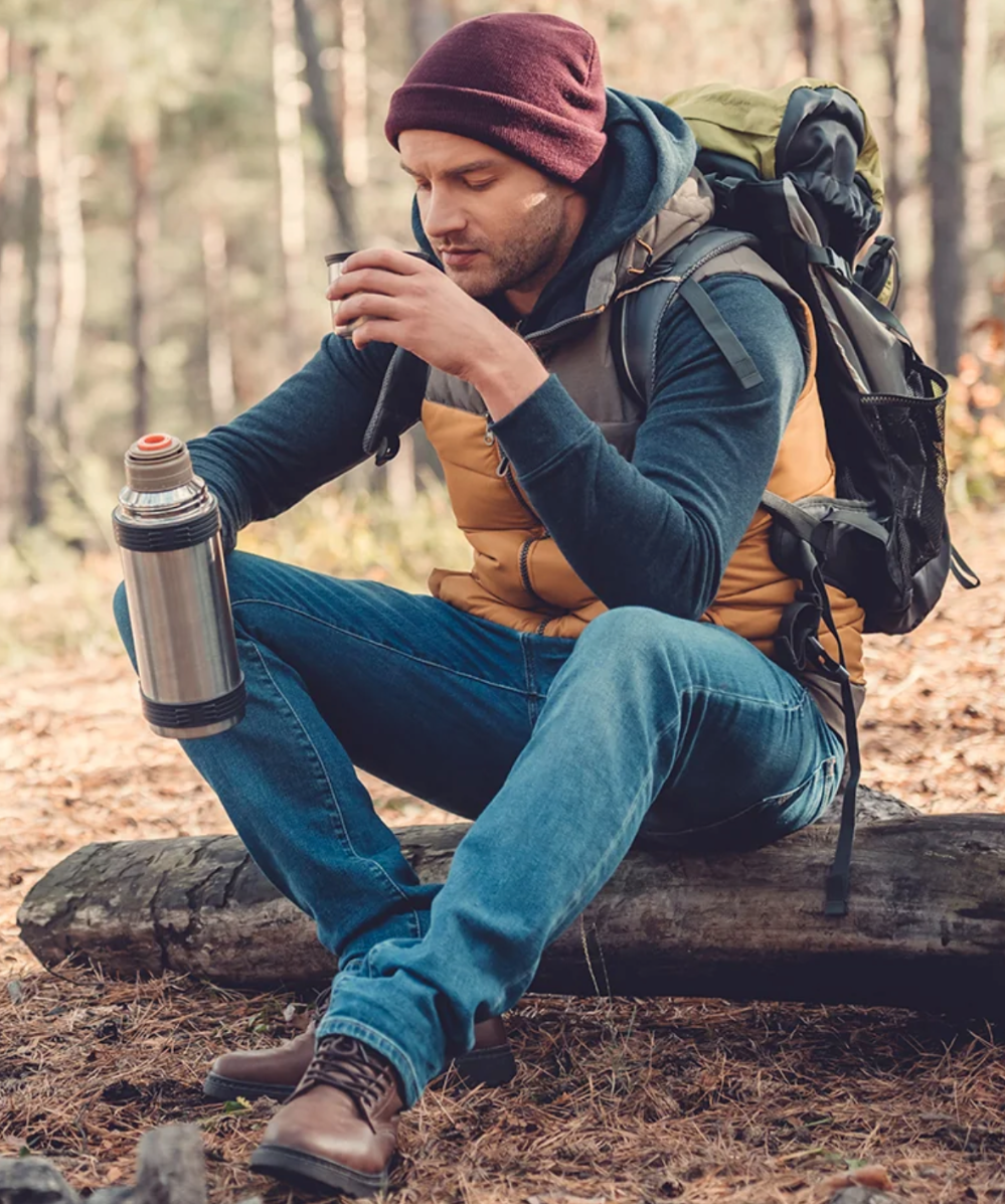 A man in American-made jeans and camping gear drinks coffee from a thermos outside.