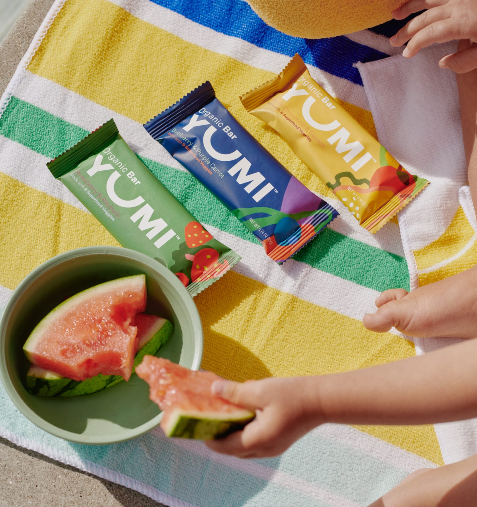 Three Yumi snack bars lie on a beach towel next to a bowl of watermelon slices.