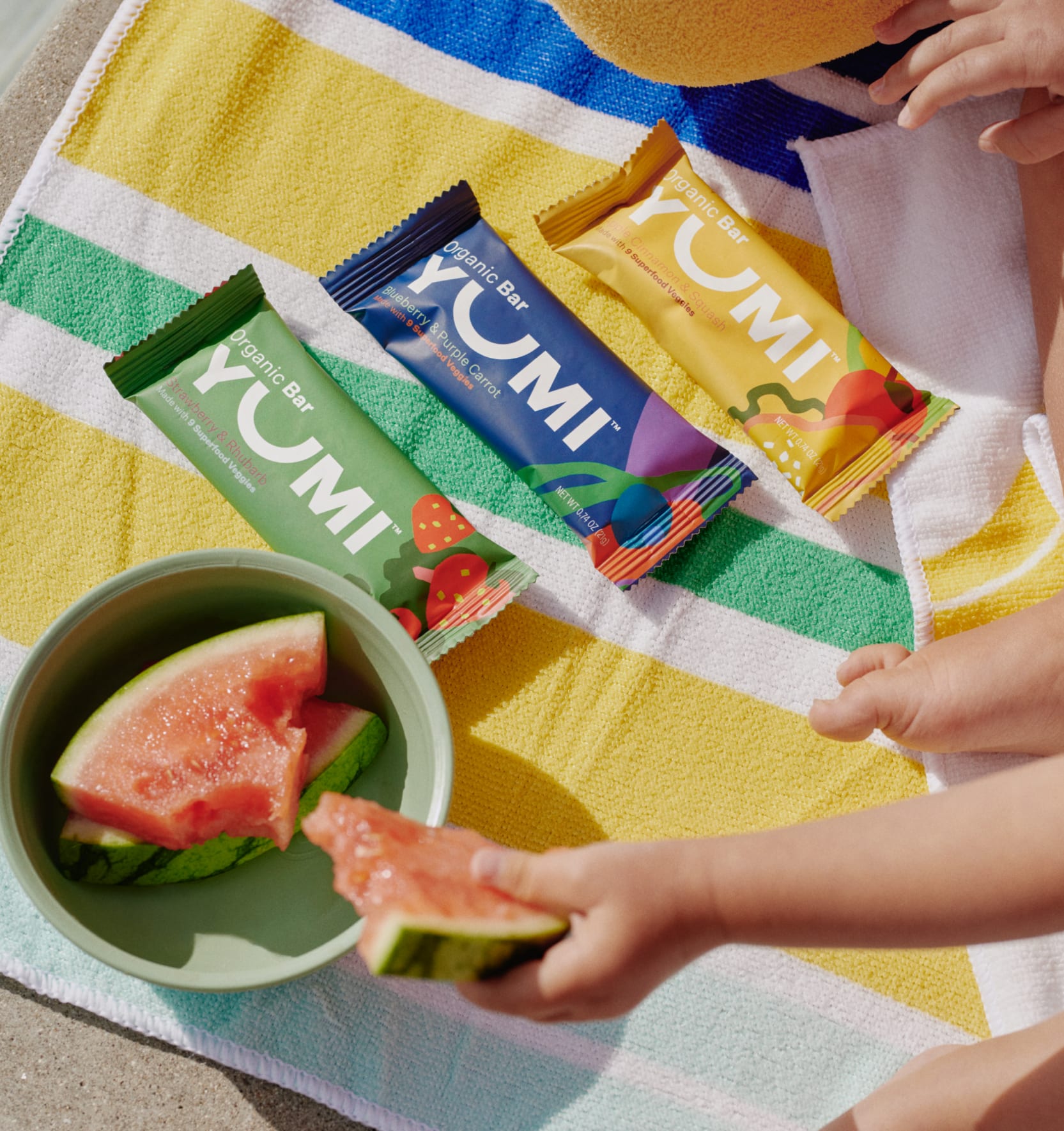 Three Yumi snack bars lie on a beach towel next to a bowl of watermelon slices. 