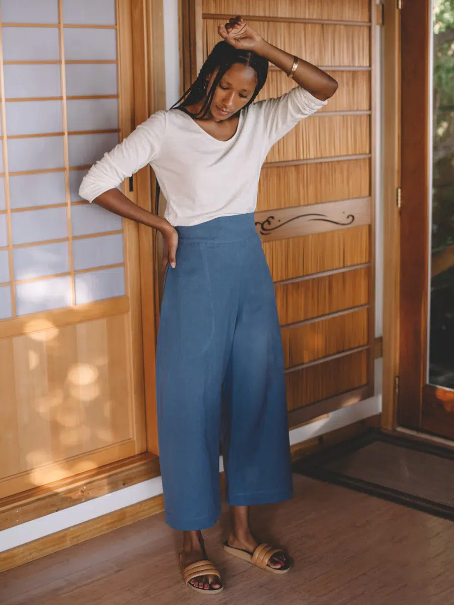 A model in indigo pants and white long sleeve shirt and slides. 