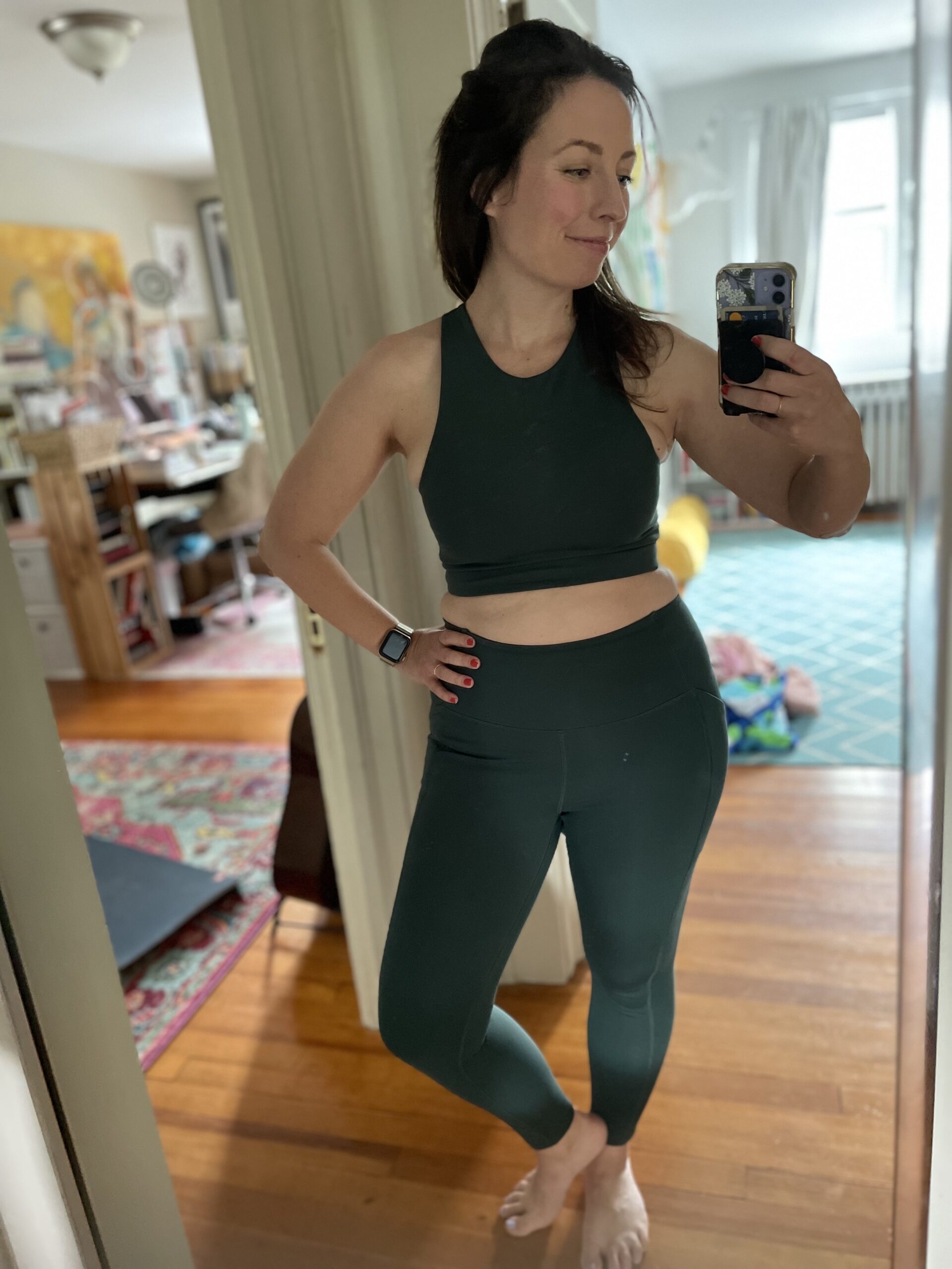 A woman takes a photo of herself wearing a sports bra and leggings in a full length mirror. 