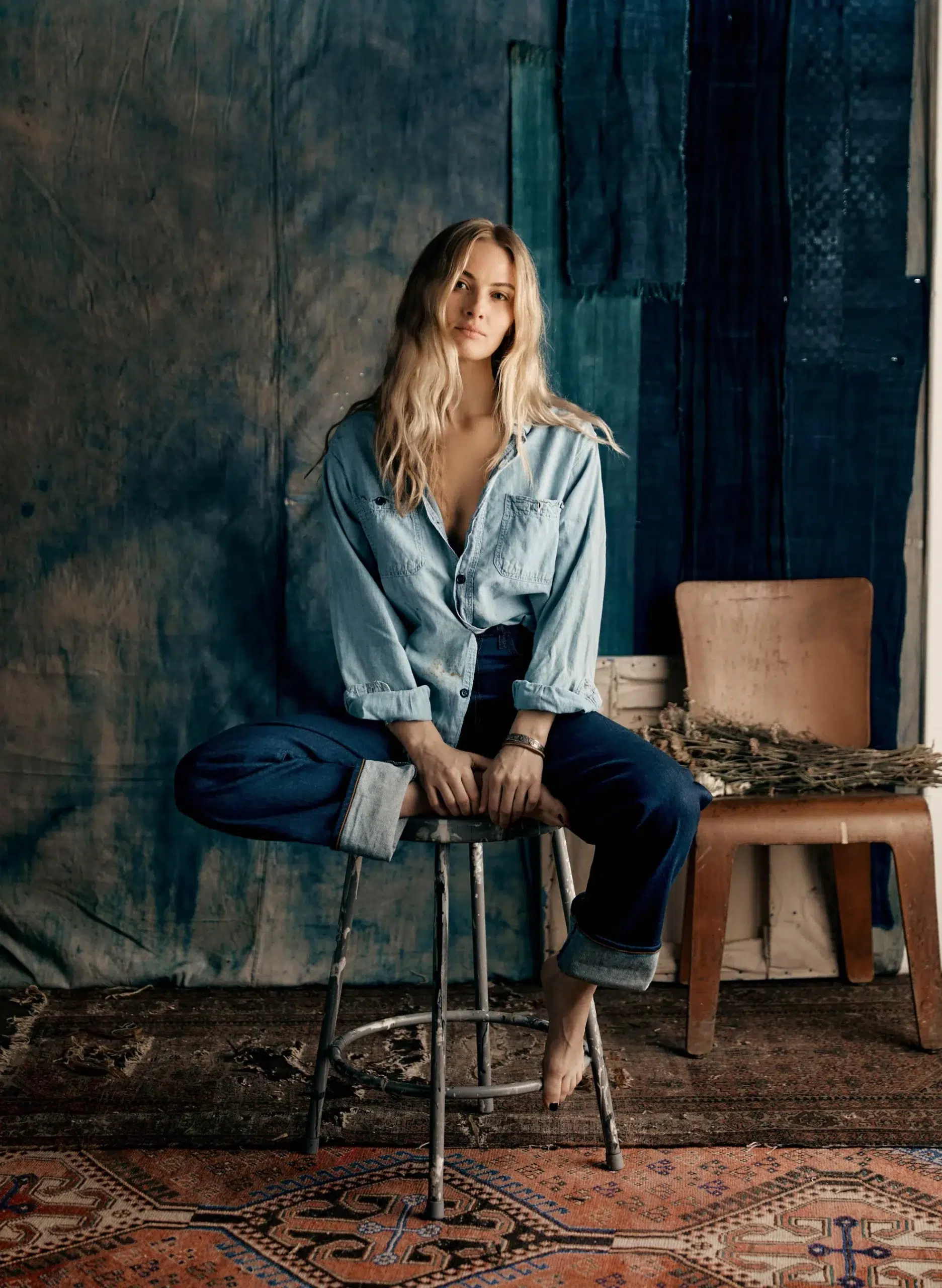 A model on a stool wearing dark American-made jeans with cuffed legs and a denim button down. 