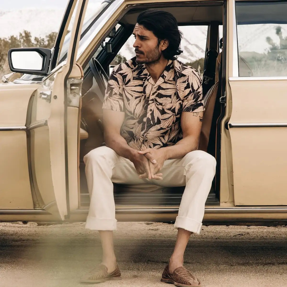 Model in a printed palm-leaf button down tee sits in an open care with his arms resting on his knees.
