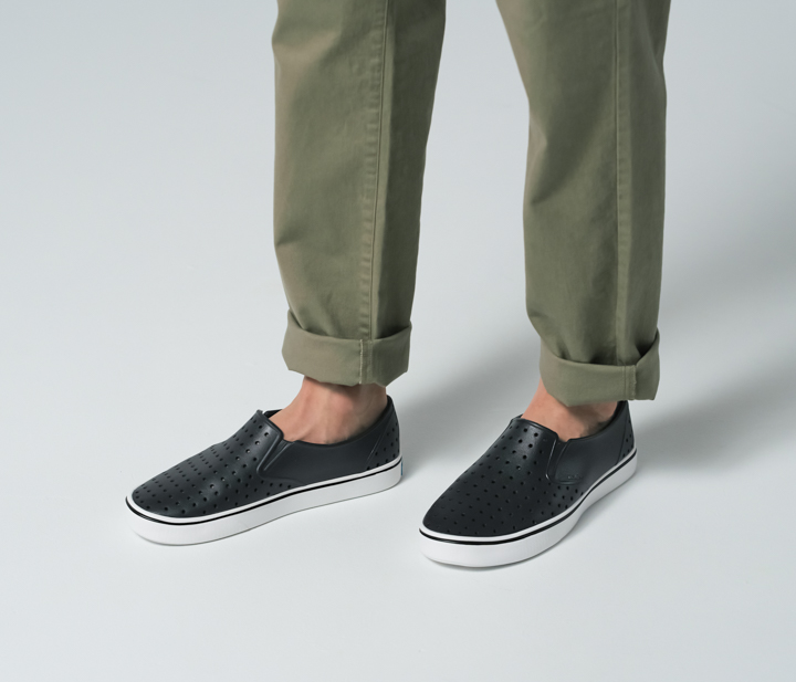 A man's legs in green pants with the cuffs rolled up wearing black slip on style Native shoes. 
