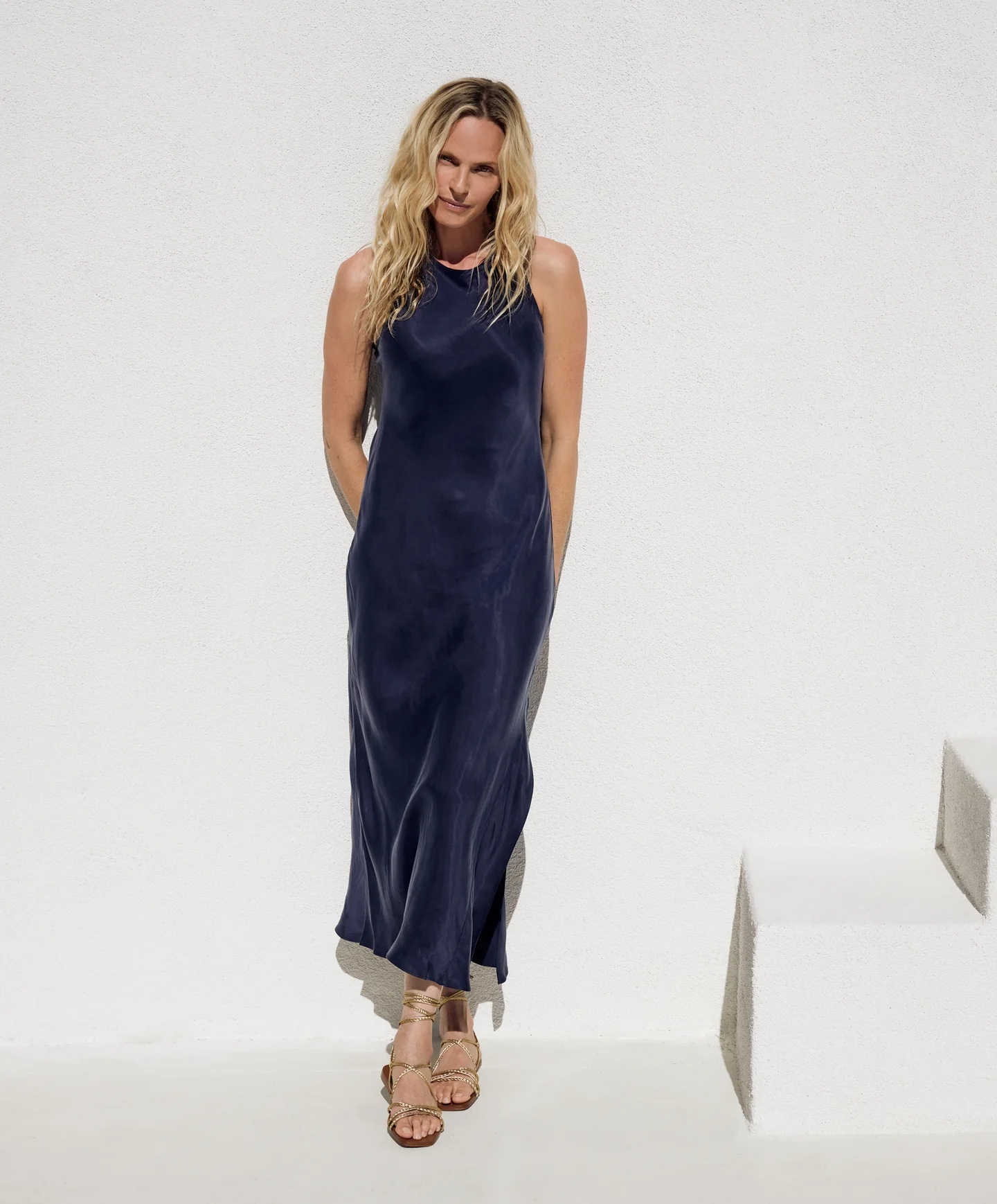 A model in a navy slip dress leans against a white wall.