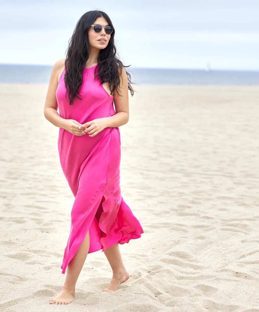 A model in a hot pink slip dress and sunglasses walks along the beach.