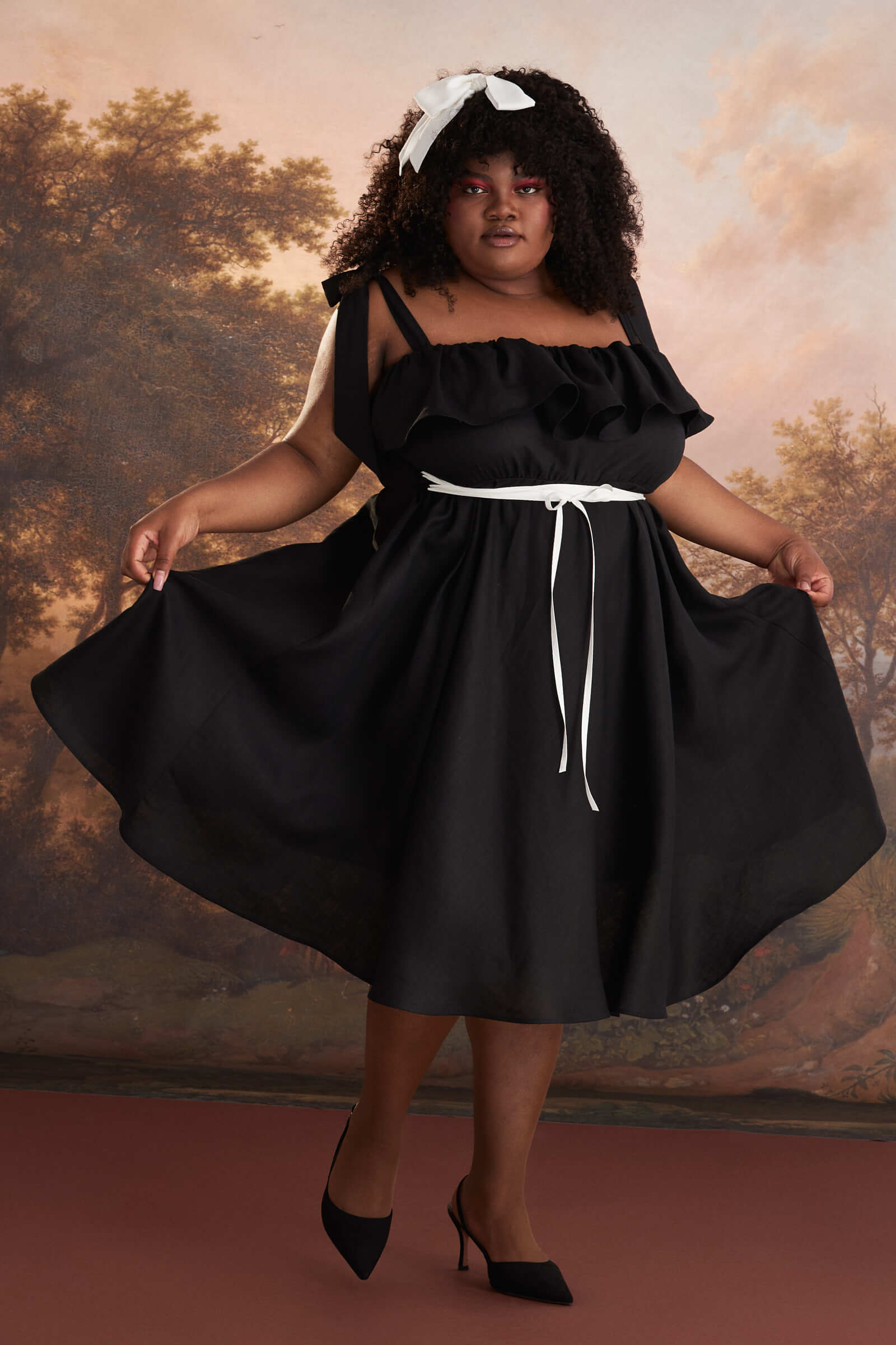 A model in front of a scenic backdrop wears a black dress with white ribbon around her waist and in her hair. 