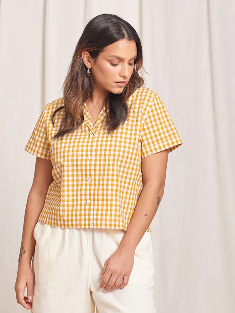 woman with long brown hair looks down, wearing a gingham checked yellow short sleeve button down
