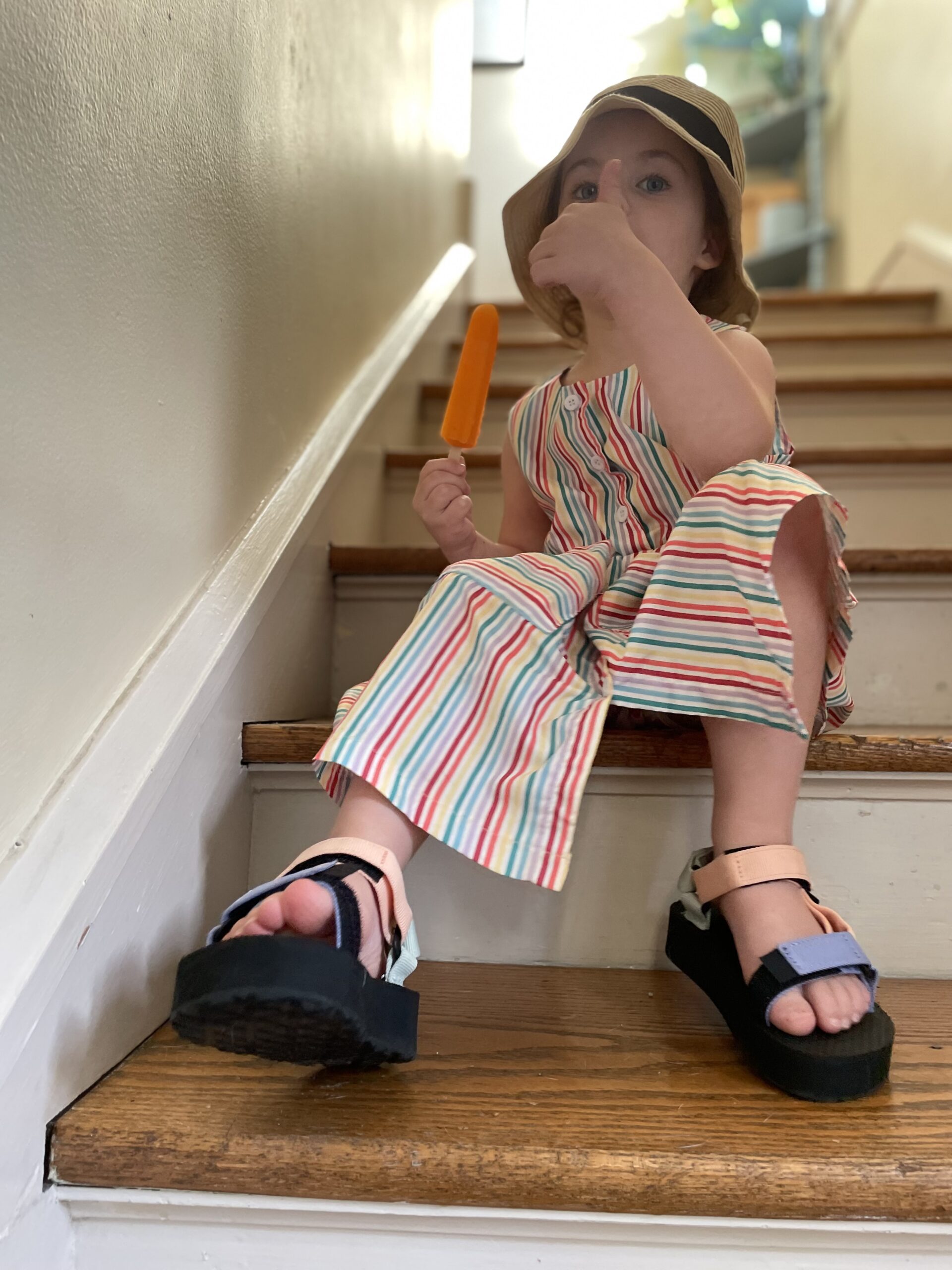 A little girl in a dress, Tevas,  and a bucket hat makes a thumbs up.