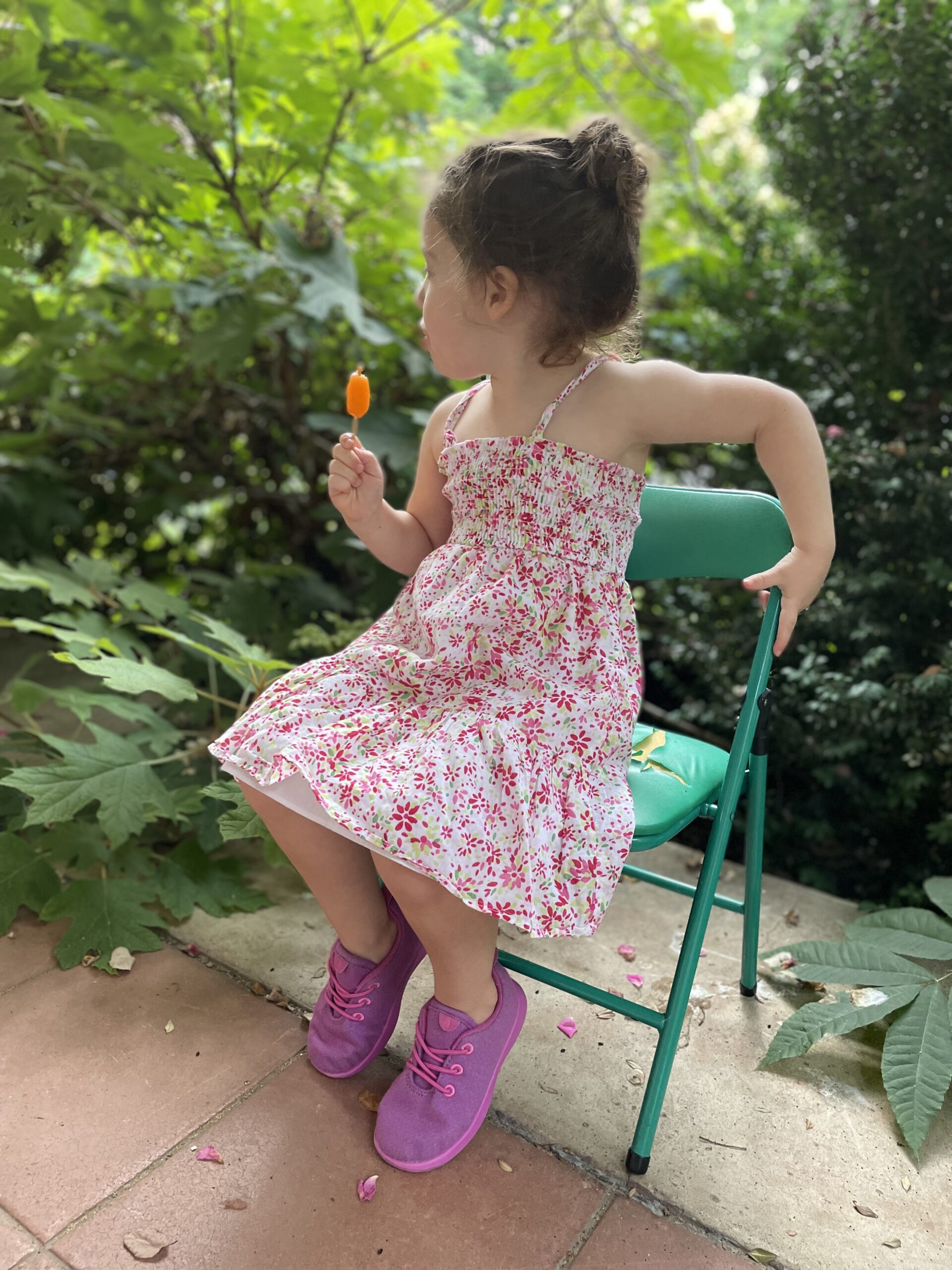 A little girl in a dress sits outside on a green chair wearing pink Allbirds wool runners.