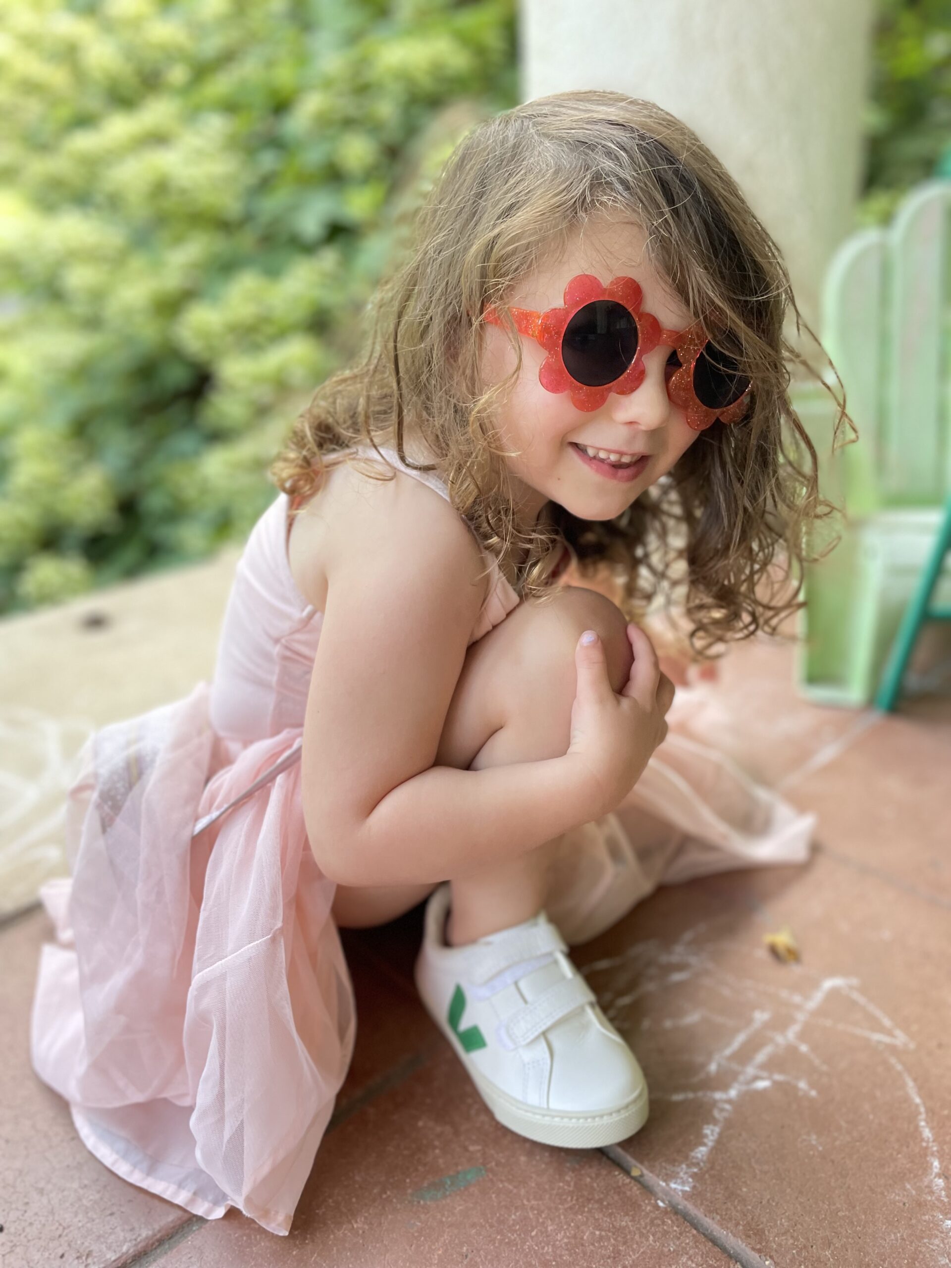 A little kid crouching in a pink dress, sunglasses, and Veja sneakers.