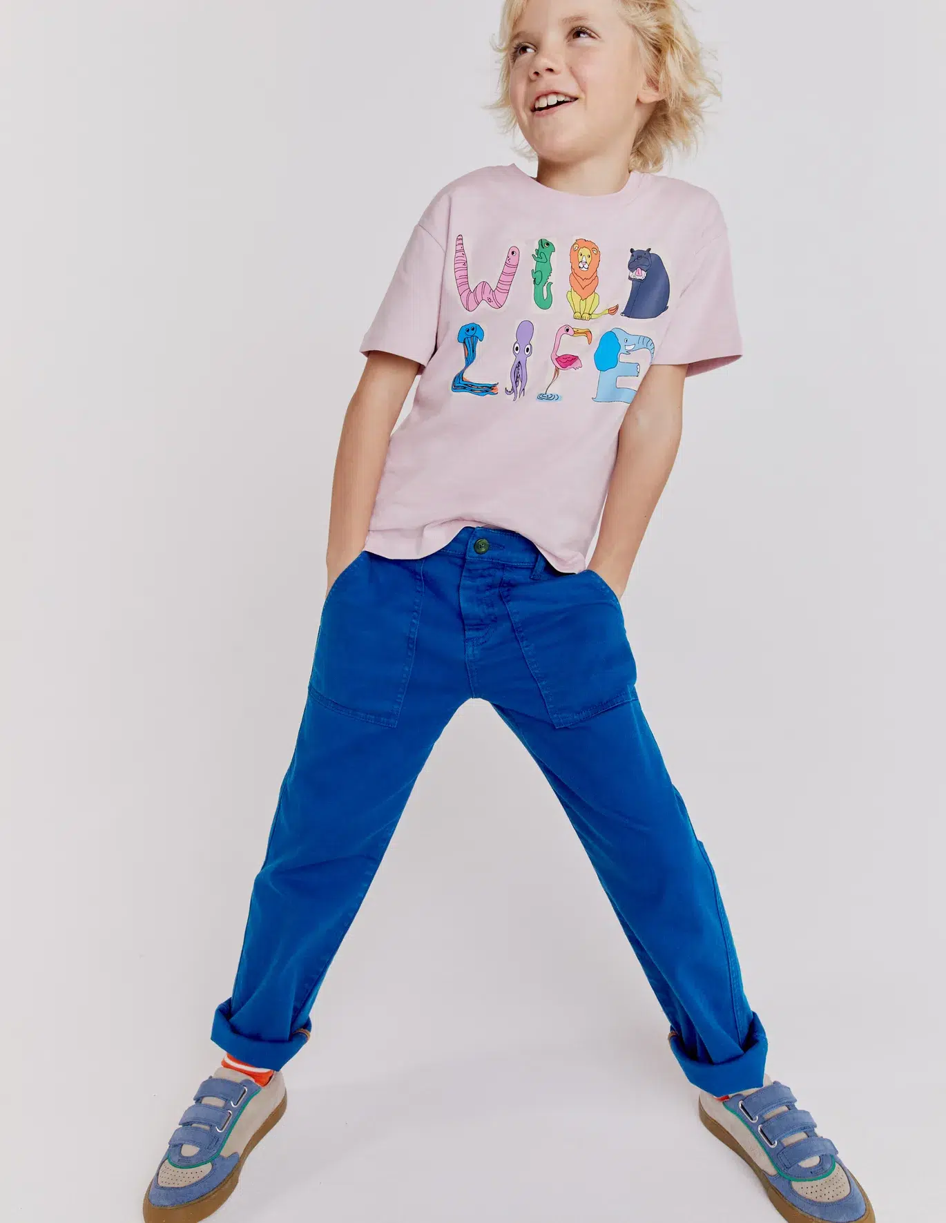 A boy in a graphic tee and blue pants has his hands in his pockets and smiles. 