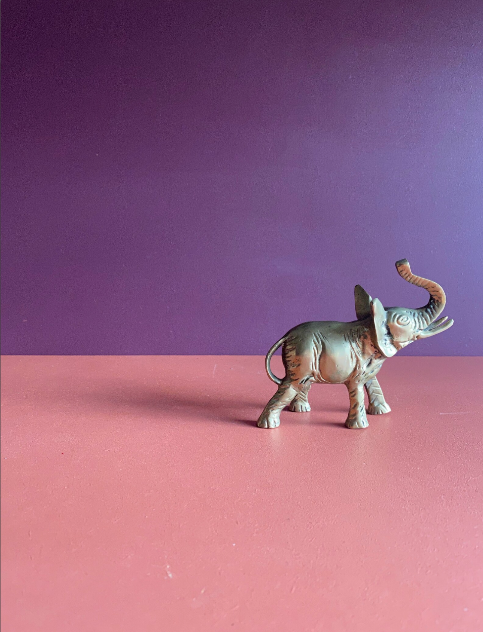 A brass elephant figurine on a pink surface against a purple wall. 