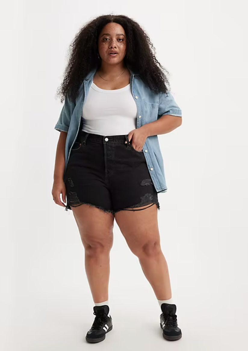 A plus size model in black cutoffs and a denim button down open over a white tank. 