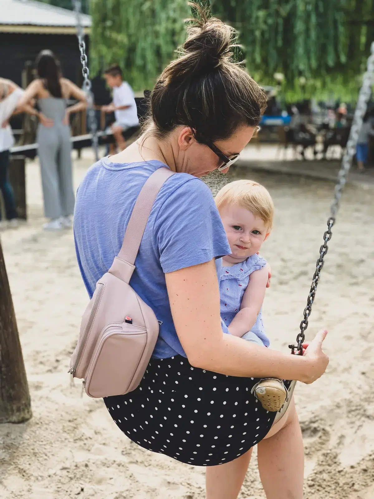 A woman holds a baby on a swing while wearing Kibou's Vegan Leather Bag across her back. 