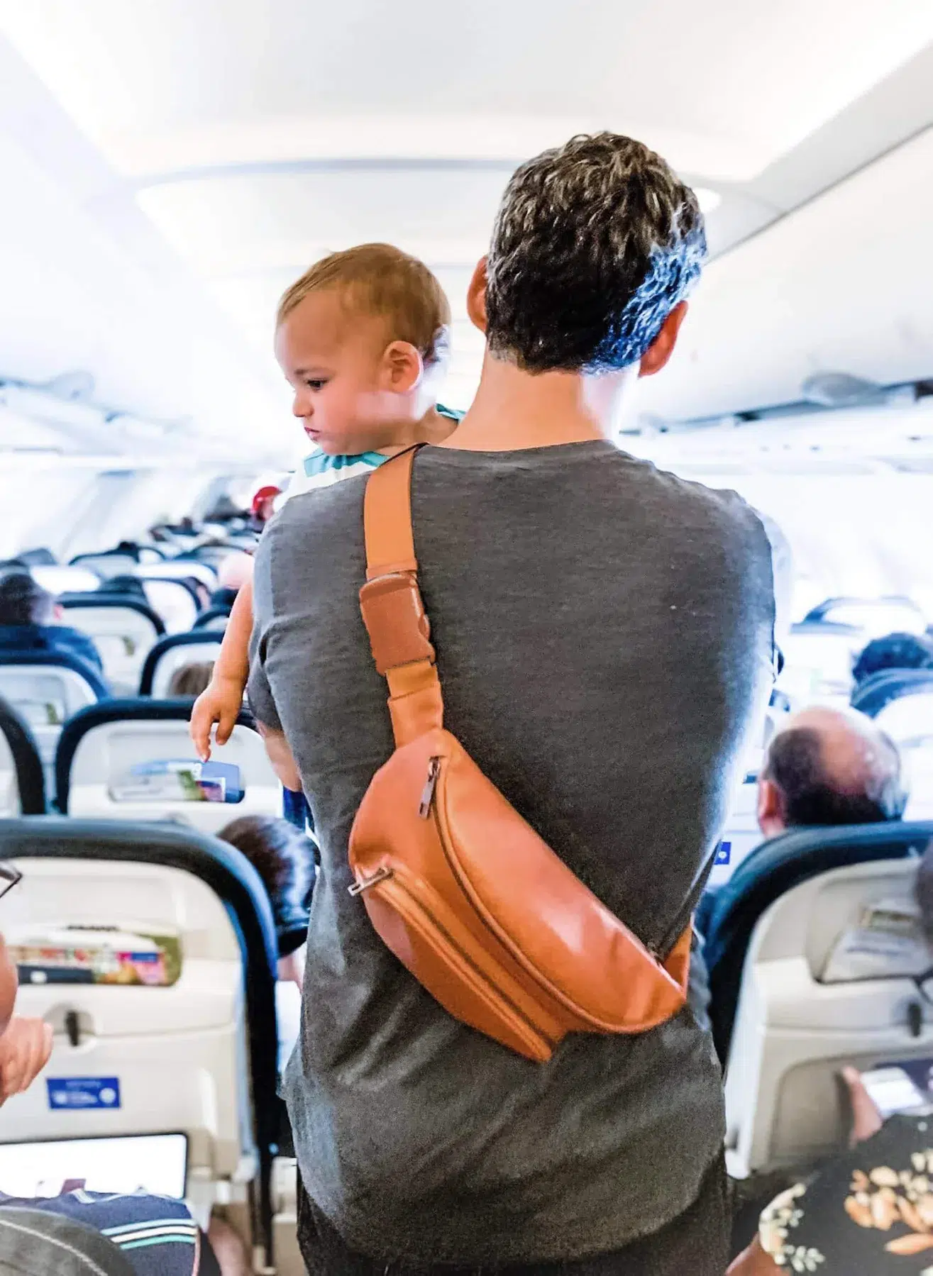A man carries a baby down a plane aisle with a Kibou Vegan Leather Bag across his back. 