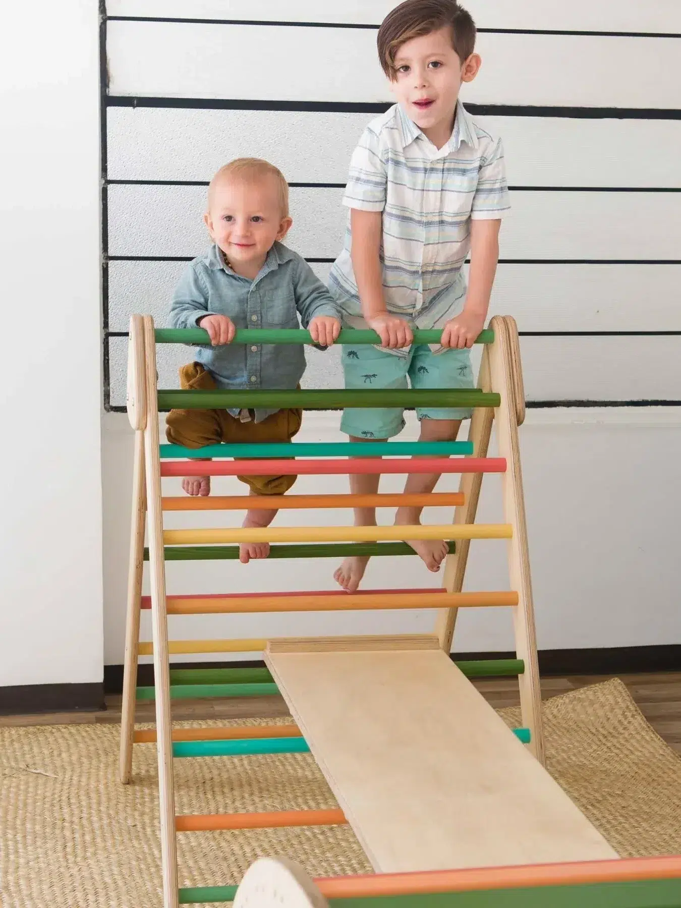 A baby and young boy stand on the rungs of a Pikler triangle. 