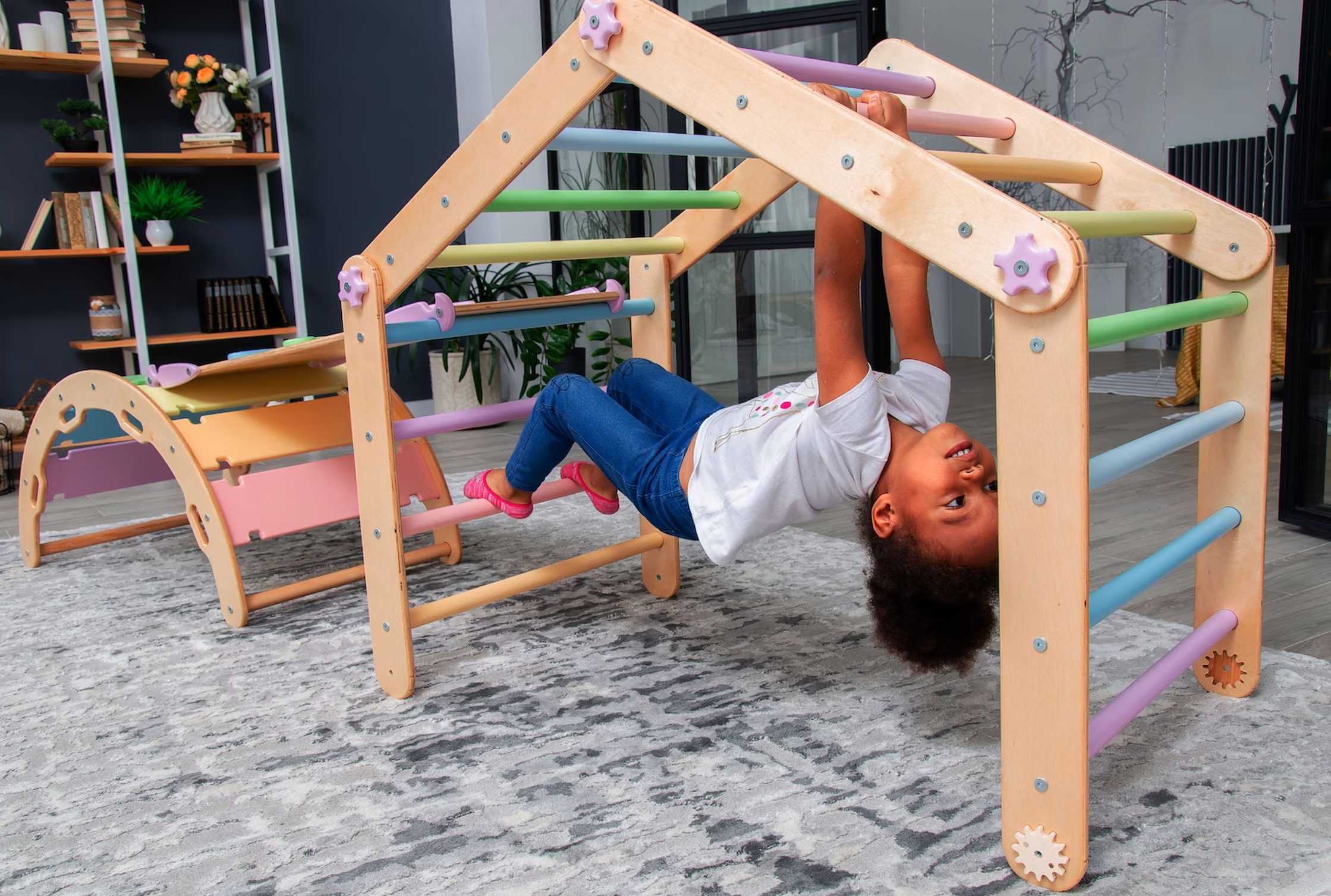 A little girl hangs on a transforming climbing triangle in the shape of a house.