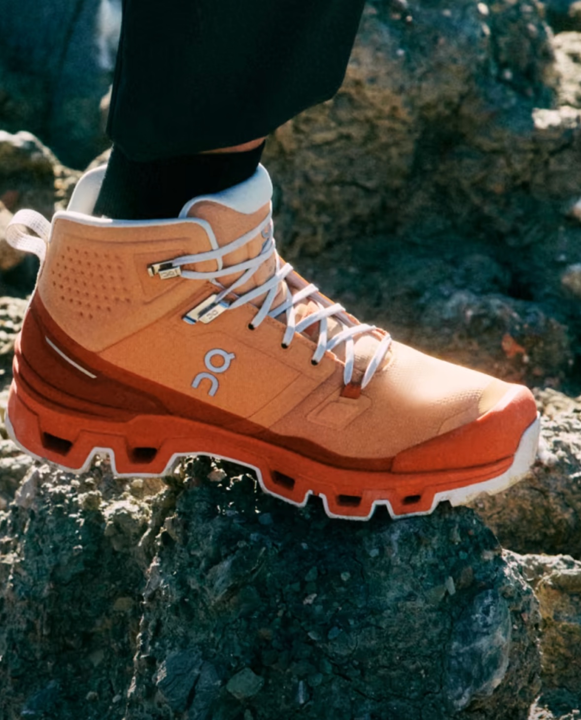A model wearing an orange On hiking boot stepping over rocks.