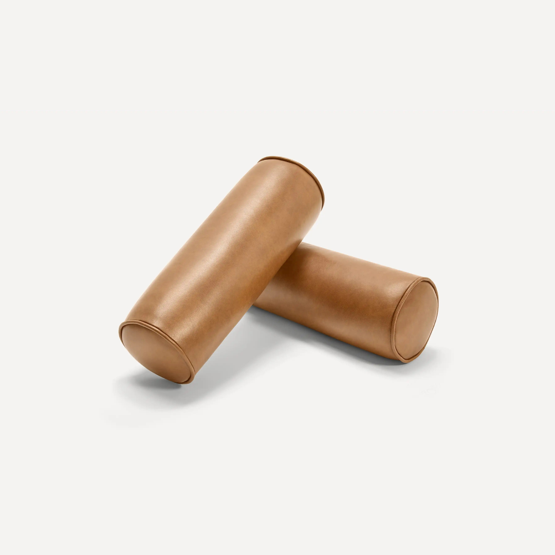 Two cylindrical brown leather bolster pillows are arranged on a plain white background, with one pillow resting slightly on top of the other.