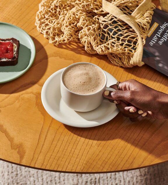 A hand holding a white mug of brewed Clevr Blends coffee alternative, set on a wooden table.
