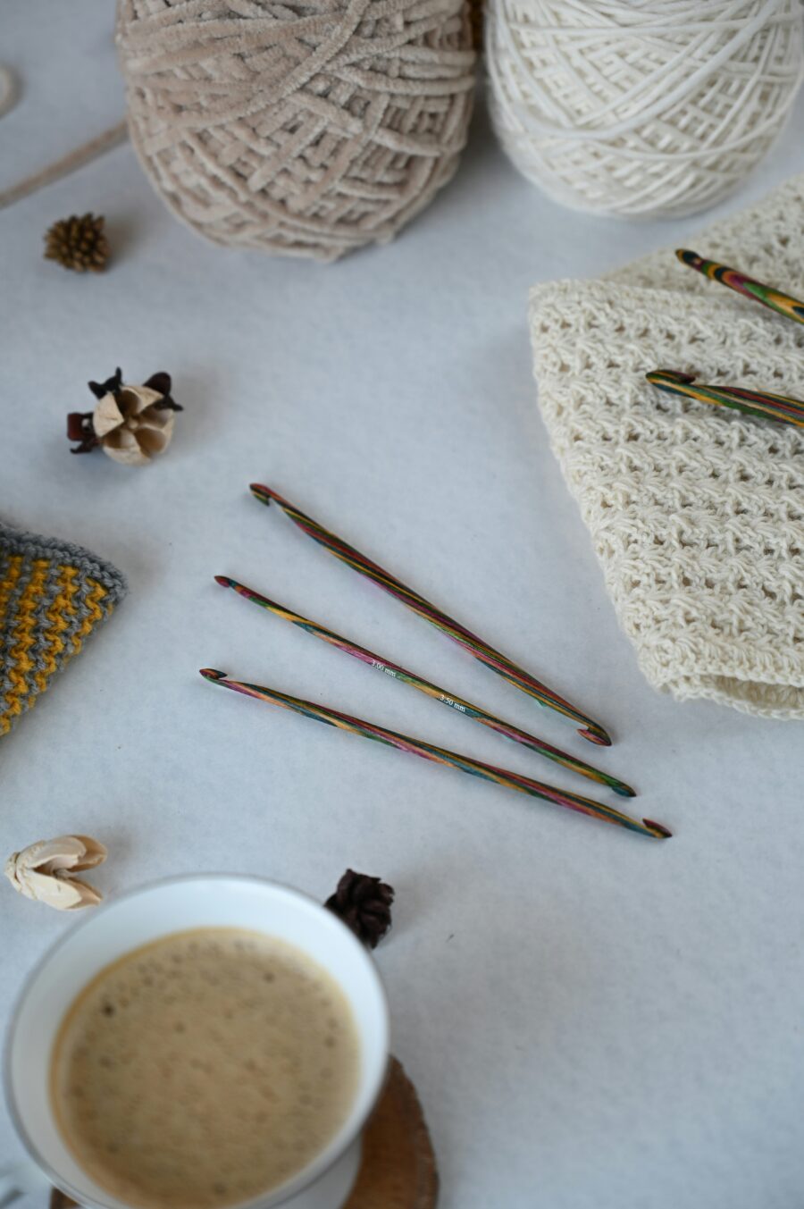 Three colorful crochet hooks, balls of yarn, crocheted fabric swatches, natural decorations, and a cup of coffee on a white surface.