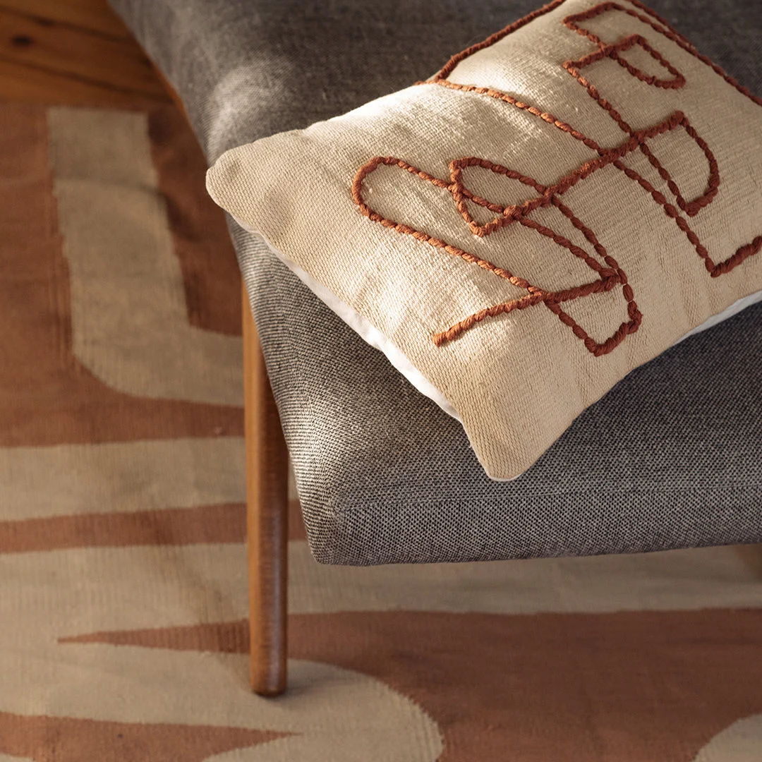 A beige pillow with abstract brown embroidery rests on a gray chair with wooden legs, next to a patterned beige and brown rug.