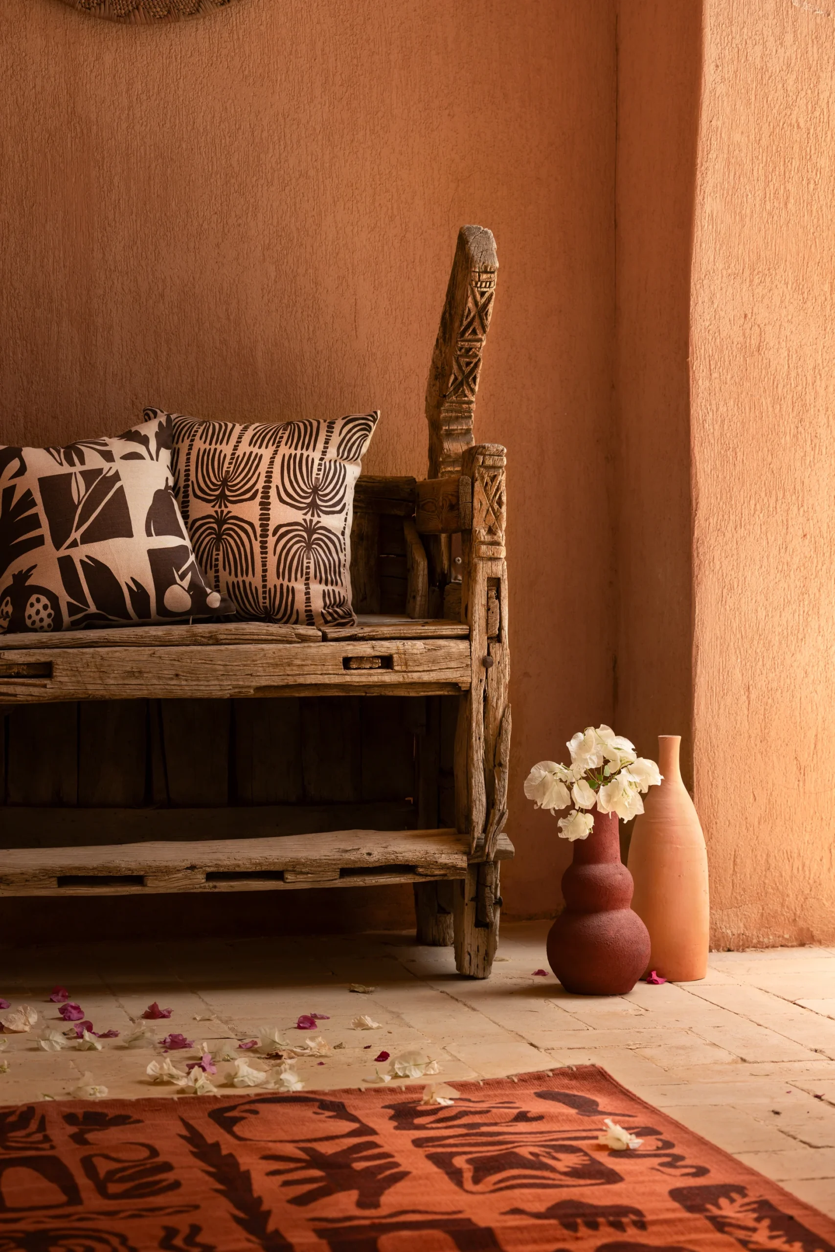 Wooden bench with patterned cushions next to a terracotta vase with white flowers, a beige vase, and scattered flower petals on a tiled floor in a sunlit room with peach-colored walls.