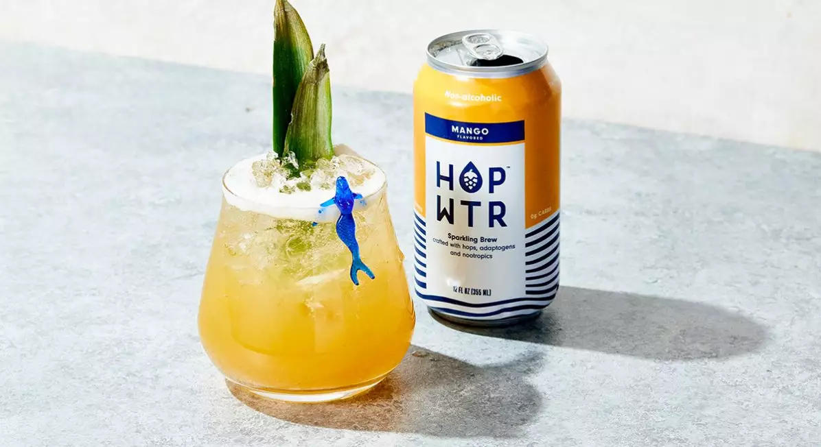 A glass of the product in a garnished glass next to a can of mango Hop Wtr. 