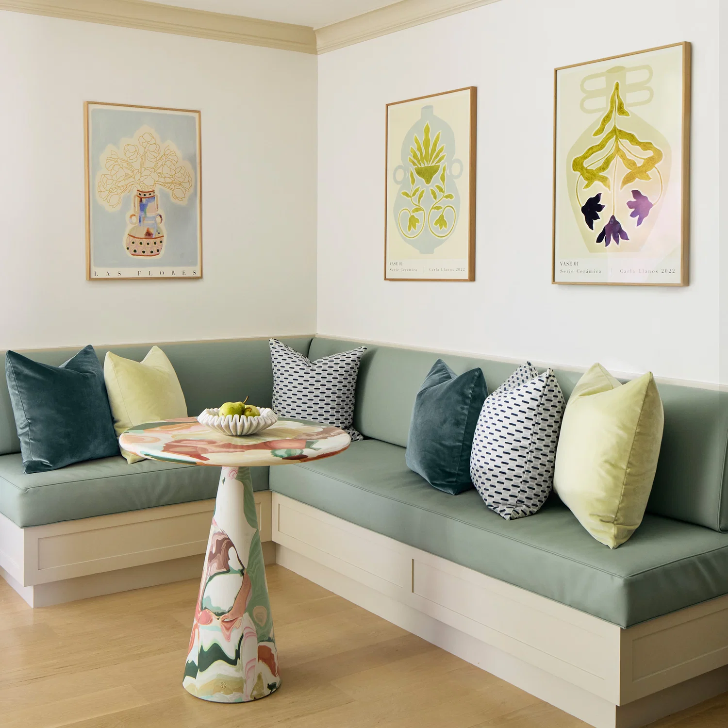 A corner dining nook with sage green bench seating, patterned throw pillows, a round table with a colorful base, and three framed botanical prints on the wall.
