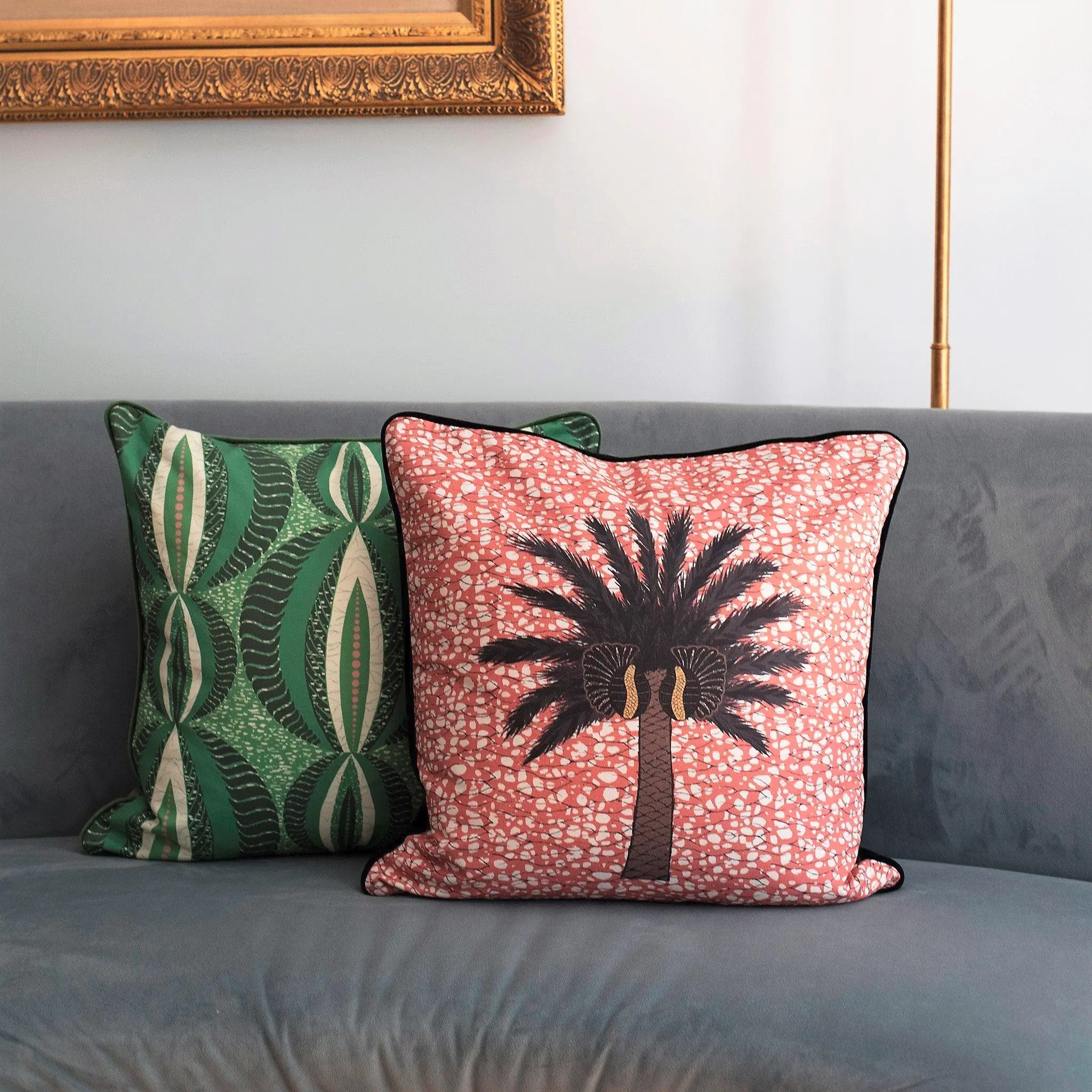 Two patterned pillows, one green with a leaf design and one pink with a palm tree, sit on a gray velvet sofa below a gold-framed painting.