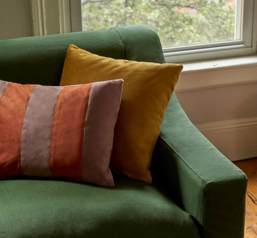 A green couch with two pillows, one solid yellow and one with orange and gray vertical stripes, sits near a window.