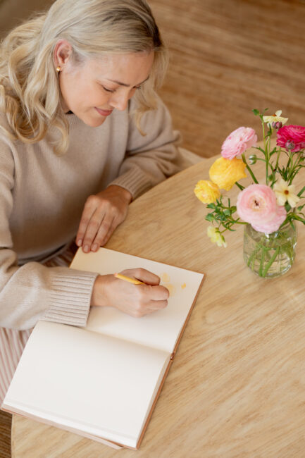 Woman drawing with a pencil in a blank notebook at a wooden table with a vase of colorful flowers nearby.