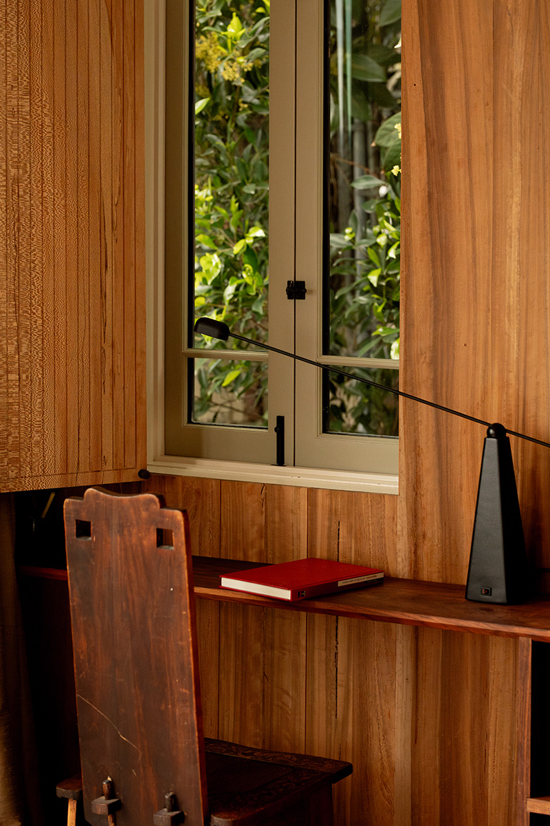 A wooden desk with a closed red notebook, a metronome, and a wooden chair sits next to a window with a view of greenery outside.