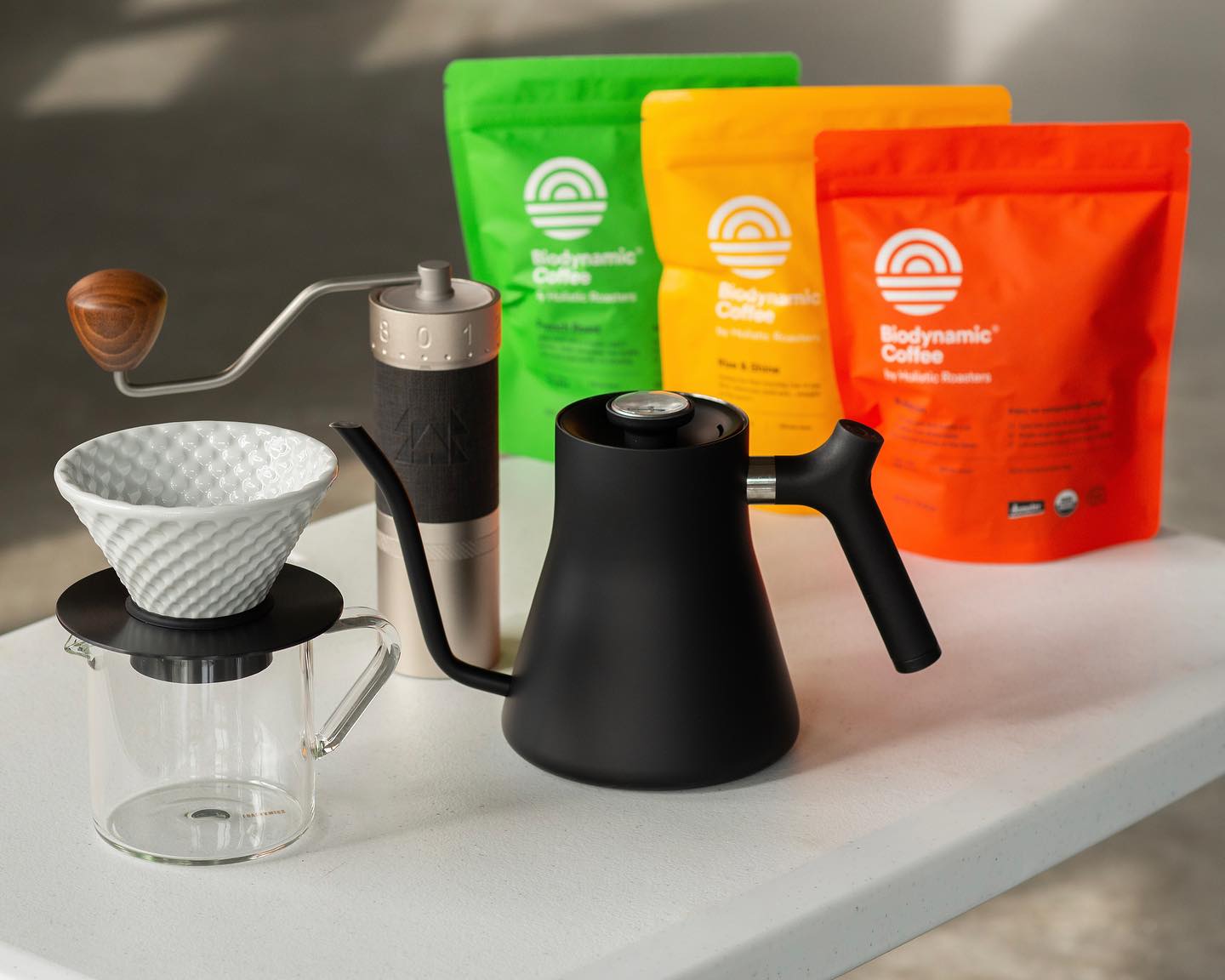 A black kettle, pour over and grinding equipment in front of a trio of bags of Holistic Roasters coffees. 