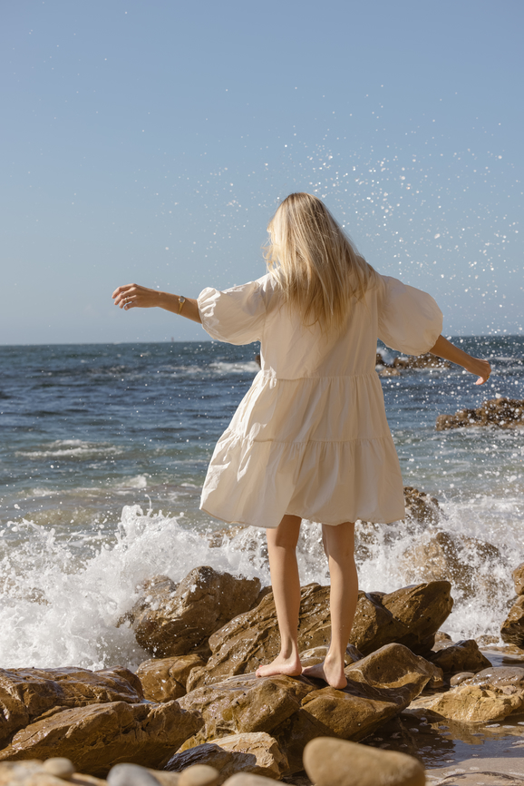 young blonde woman in white dress standing on rocks by the ocean