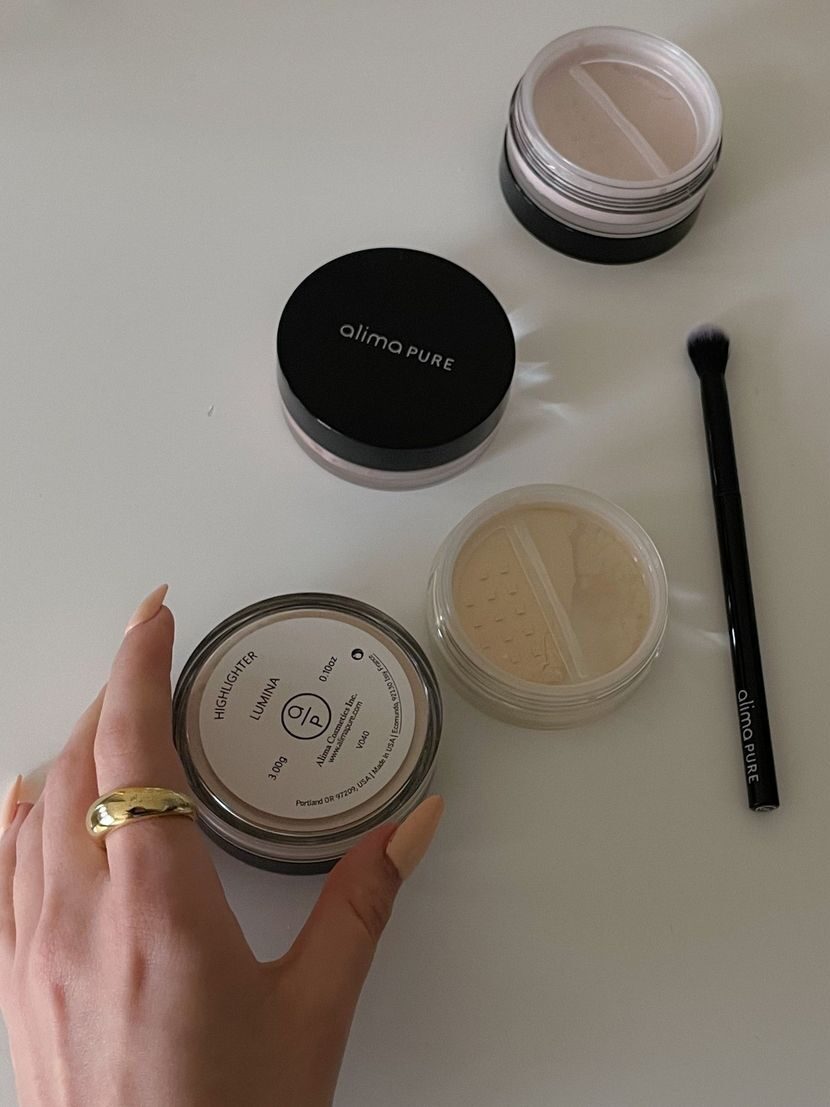 Alima Pure makeup powder containers scattered over a white table with a hand holding onto one of the powders.