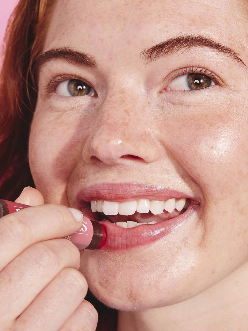 A closeup headshot of a woman with ginger hair applying a Burt's Bees lip product.