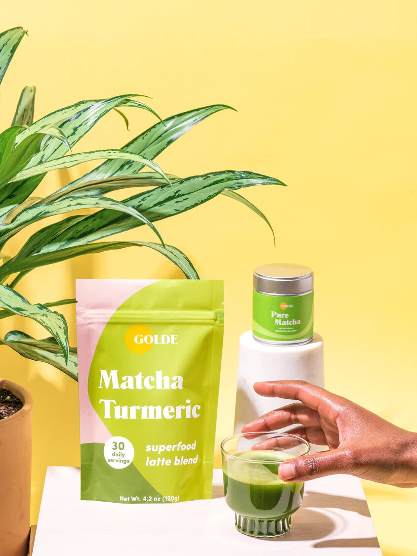 A pack of Golde Matcha Turmeric coffee alternative. To the right is a short glass filled with the blend and a hand gently holding the glass.