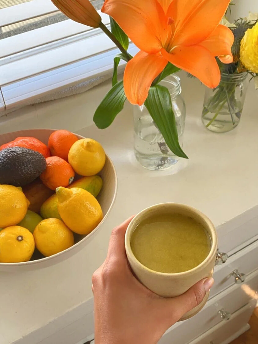 A hand holding a mug of Golde Matcha Turmeric coffee alternative over a dresser. A bowl of fruit and two vases of flowers are also on the dresser.