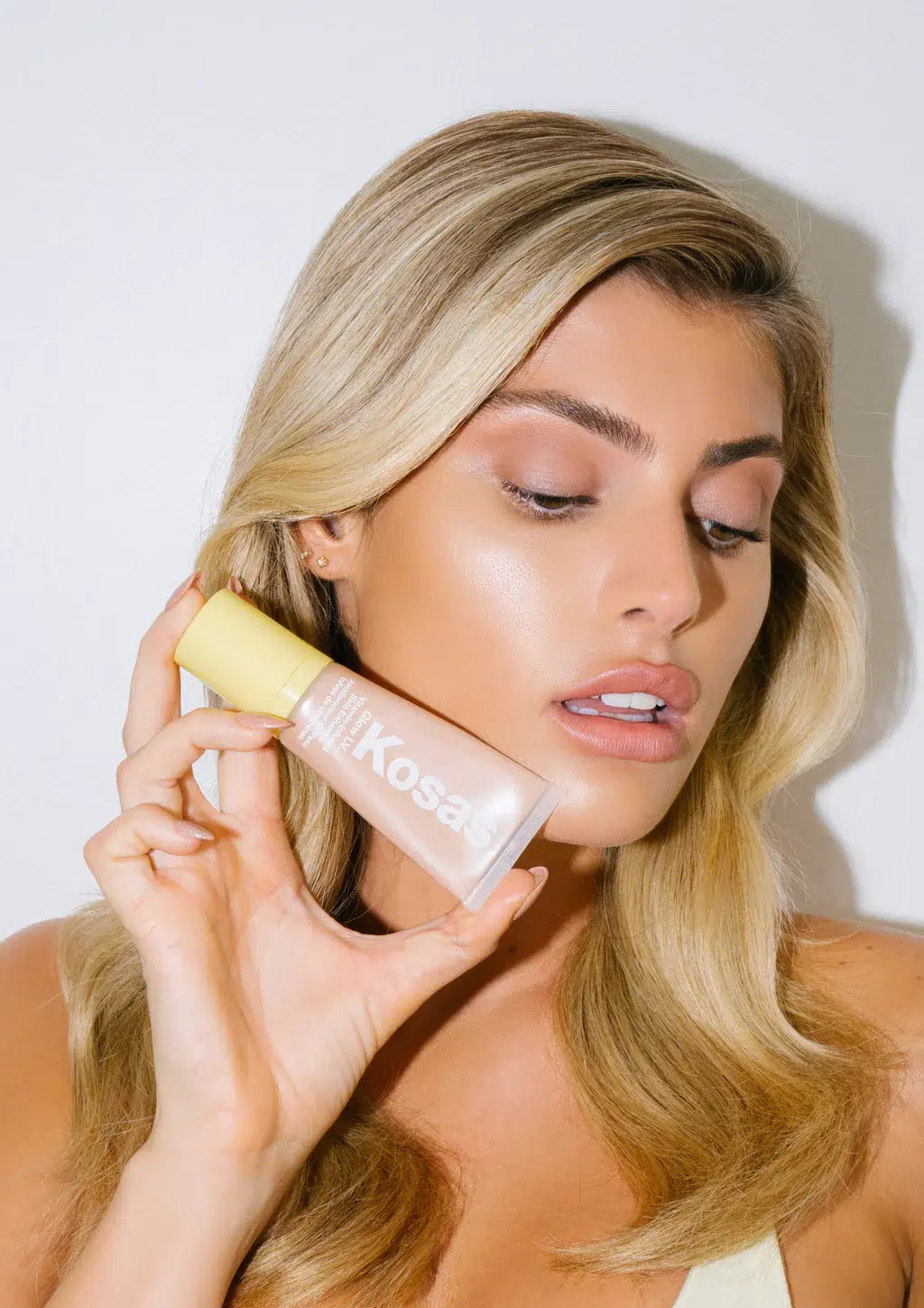 A model holds a tube of Kosas highlighter next to her cheek. 