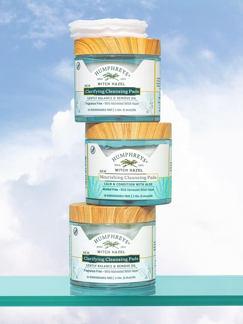 Three Humphreys Organic Witch Hazel Cleansing Pads containers stacked on top of one another on a green counter with a light blue sky and clouds in the background.
