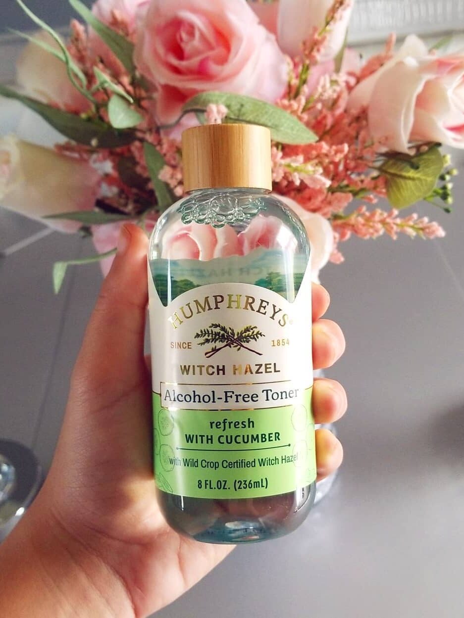 A hand holding a bottle of Humphreys Witch Hazel Spray with a bouquet of pink roses in the background.