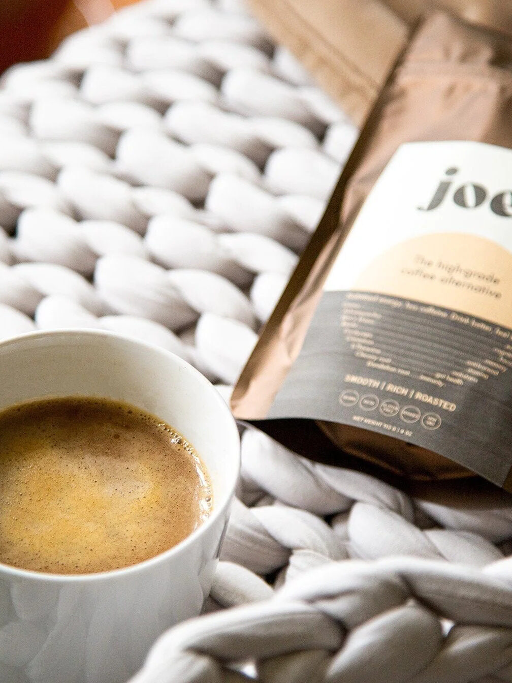 A pack of Joe'y coffee alternative laid flat with a brewed cup of the blend in front of it.