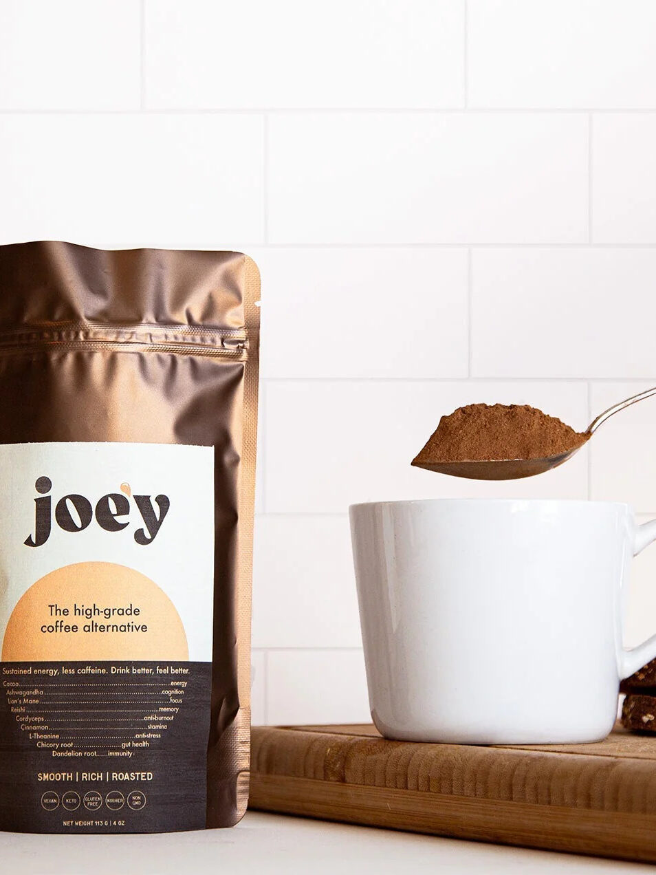 A pack of Joe'y coffee alternative to the left of a white mug. A spoon filled with the coffee alternative is about to be added to the mug.