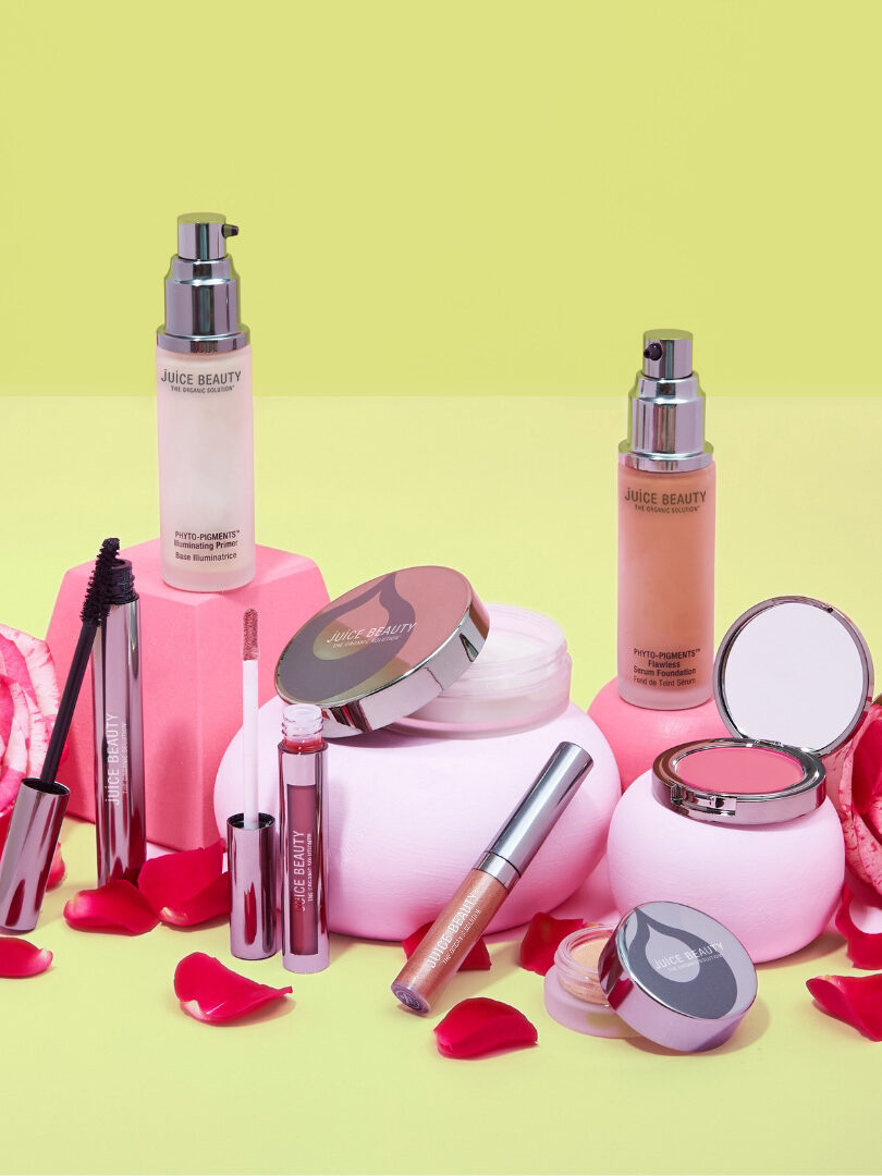 An array of Juice Beauty makeup products displayed in front of a lime green background, surrounded by pink flowers.