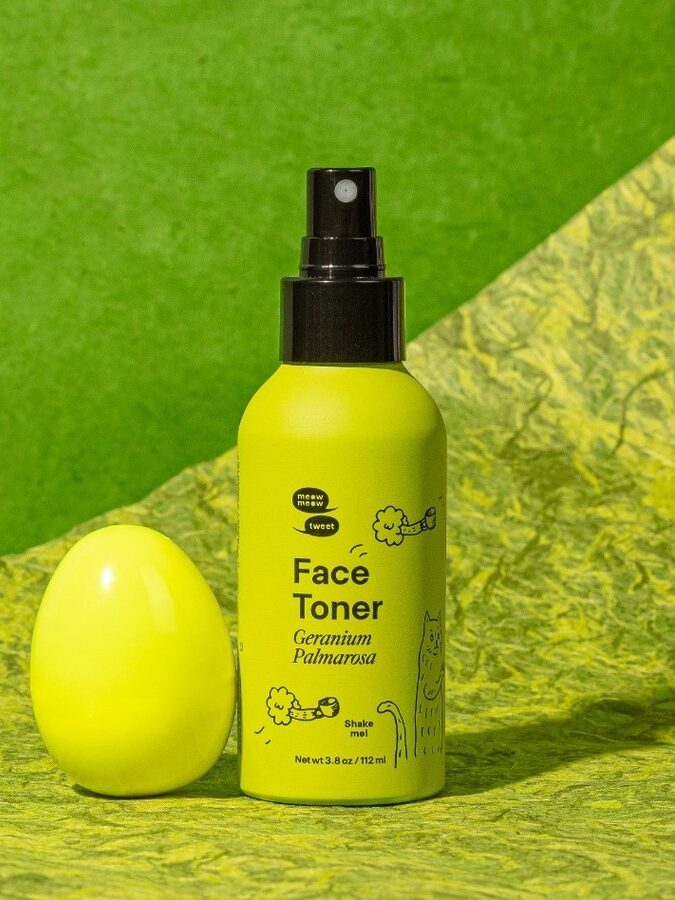 A bottle of Meow Meow Tweet's Face Toner with a green egg to the left of the bottle. The background is a two-tone marble green separated diagonally.