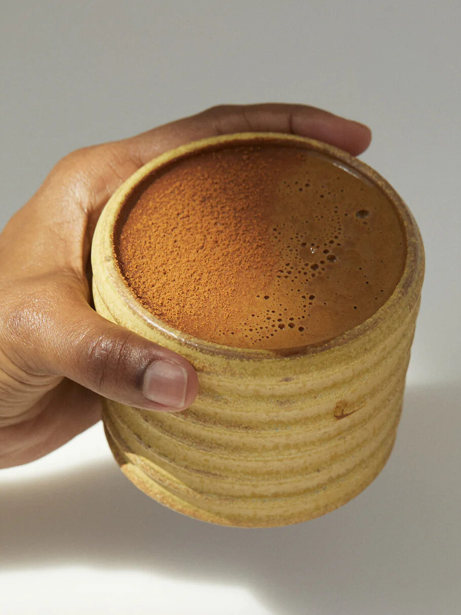 A hand holding a brewed mug of MudWtr coffee alternative in a yellow handleless mug.